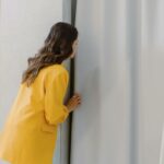 A brunette woman in a yellow blazer peeks through elegant fitting room curtains in a store.